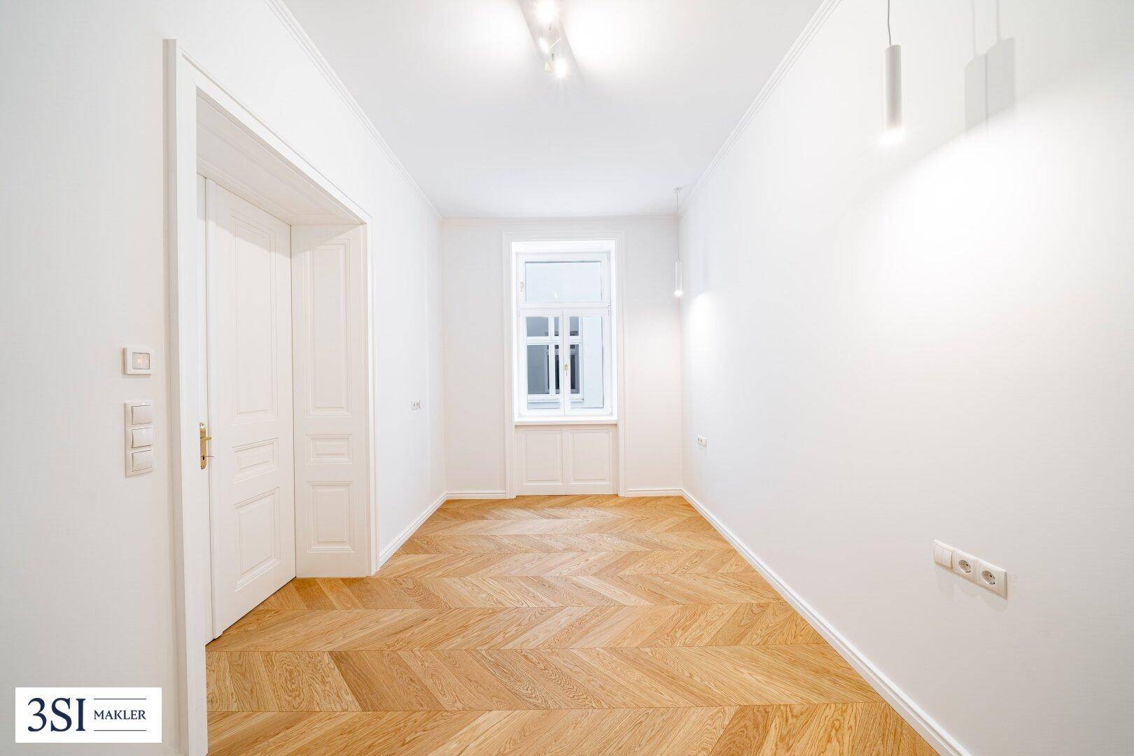 Langer, heller Raum mit Fischgrätparkett und einem Fenster, ideal als Flur oder kleines Zimmer.