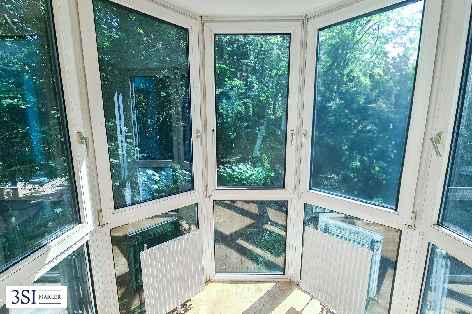 Heller Erker mit vielen Fenstern und Blick auf grüne Bäume.