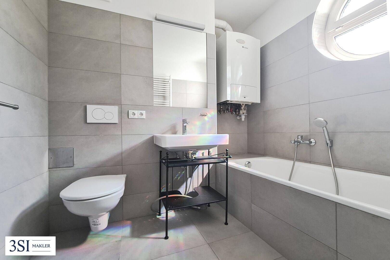 Modernes Badezimmer mit grauen Fliesen, Badewanne, wandhängendem WC und Fenster.