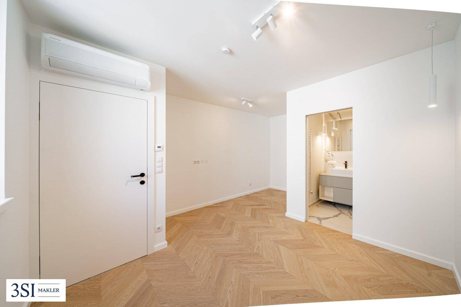 Heller Raum mit Fischgrätparkett und Blick auf einen modernen Badezimmerbereich.