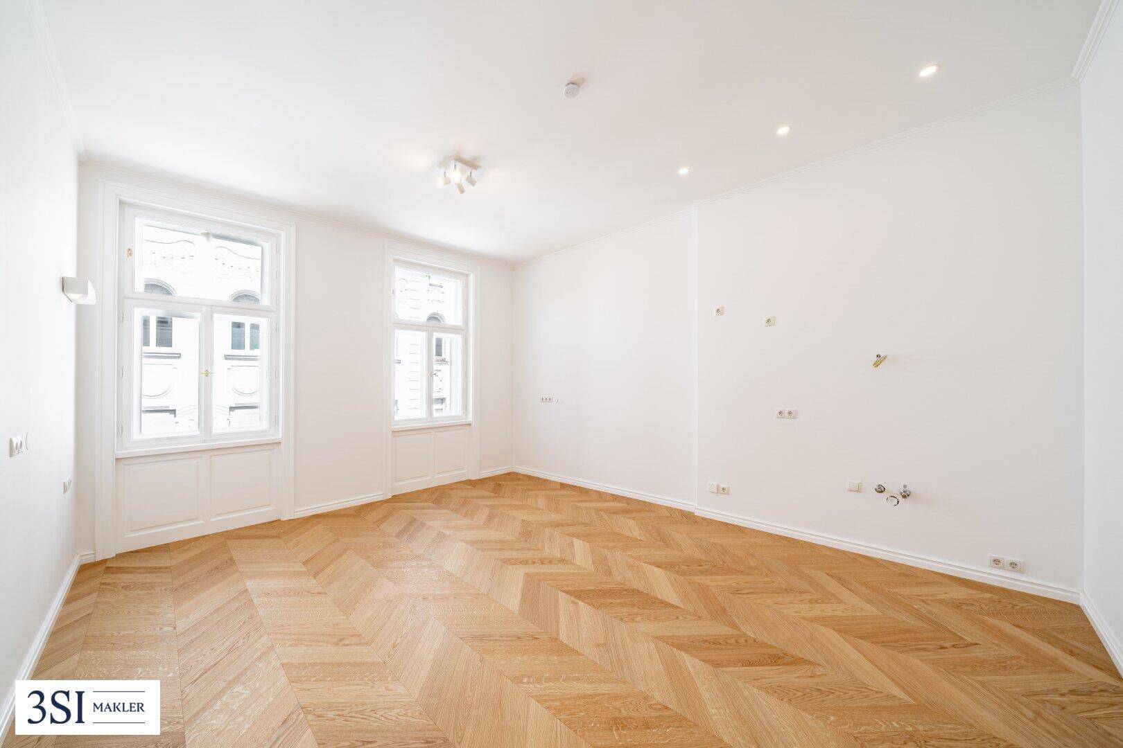 Heller, leerer Raum mit elegantem Fischgrätparkett und hohen Fenstern.