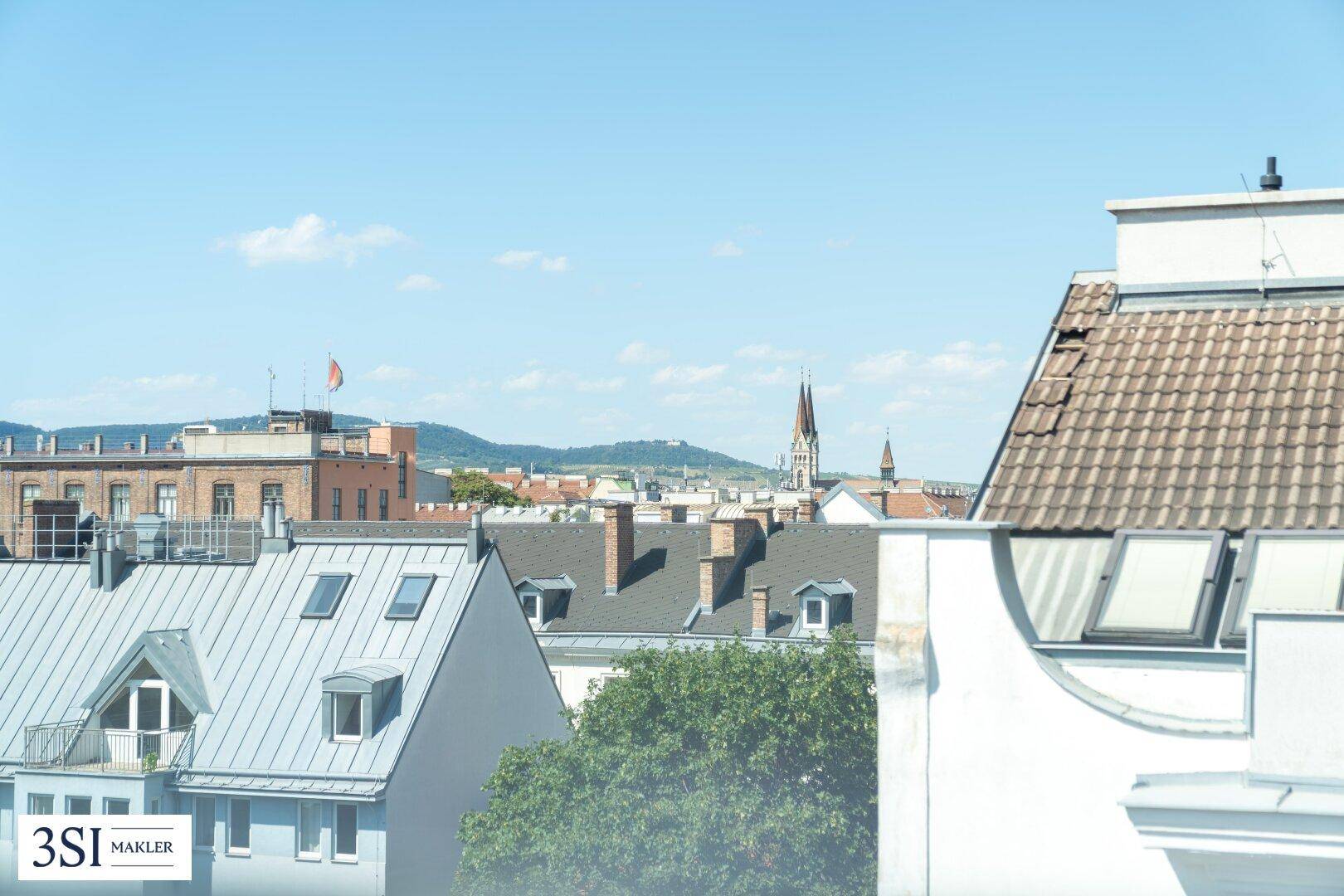 Panoramablick über die Dächer der Stadt mit Hügeln und einem Kirchturm im Hintergrund.