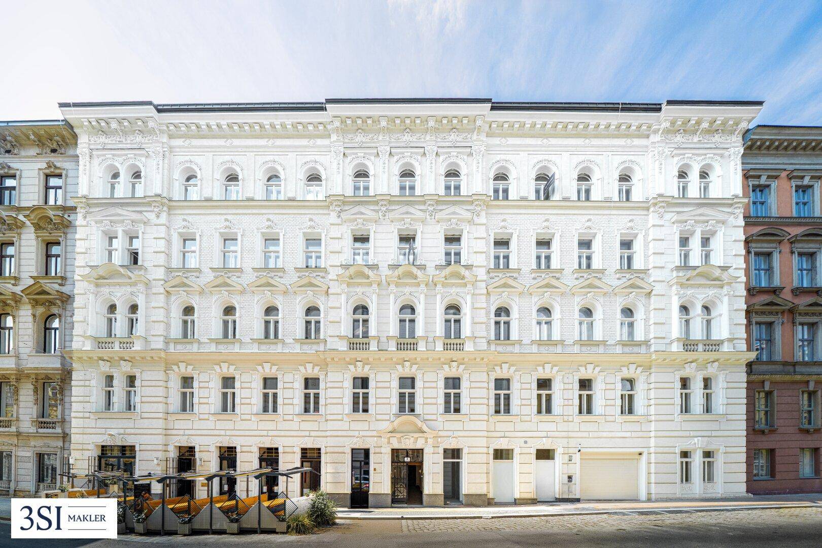 Weißes, historisches Gebäude mit reich verzierter Fassade und vielen Fenstern unter blauem Himmel.