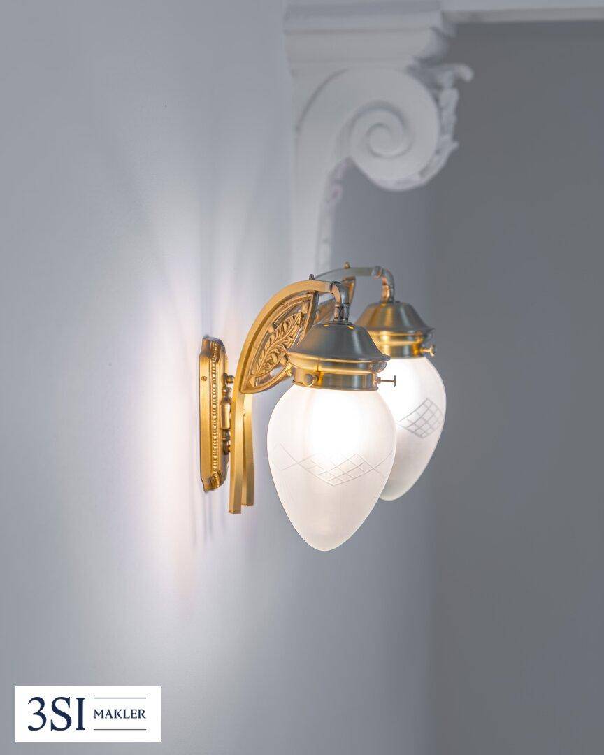 Detailaufnahme einer eleganten, goldenen Wandleuchte mit zwei milchigen Glaslampen und Stuckverzierung.