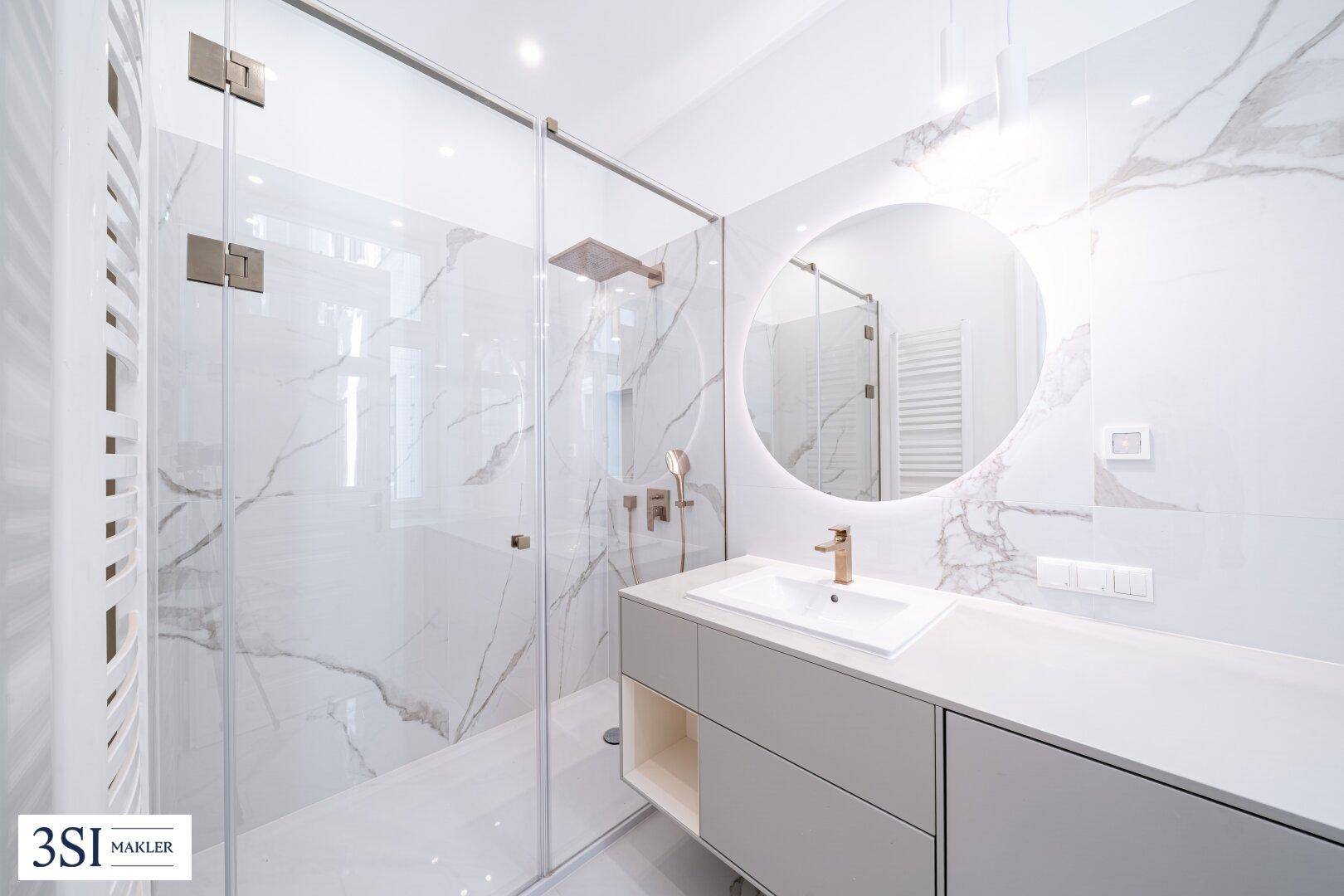 Modernes Badezimmer mit Marmorfliesen, beleuchtetem Spiegel, Einzelwaschbecken und begehbarer Dusche.