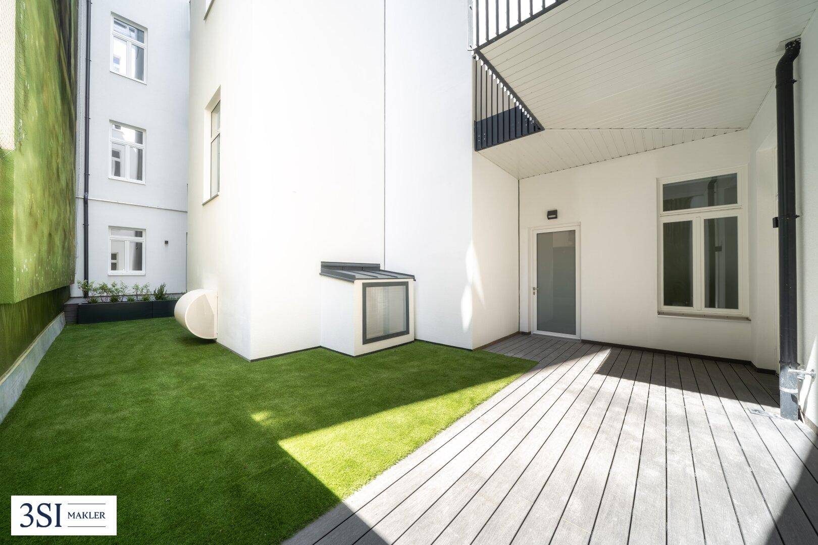 Kleiner privater Gartenbereich mit Kunstrasen und Holzterrasse, umgeben von Gebäuden.