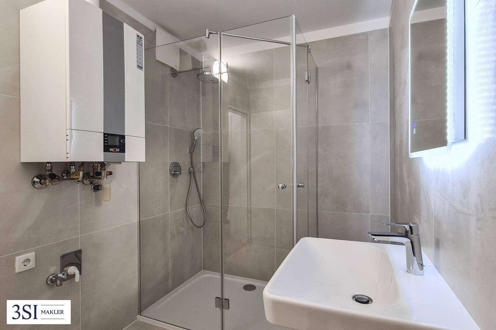 Modernes Badezimmer mit grauen Fliesen, Duschkabine, Waschtisch und beleuchtetem Spiegel.