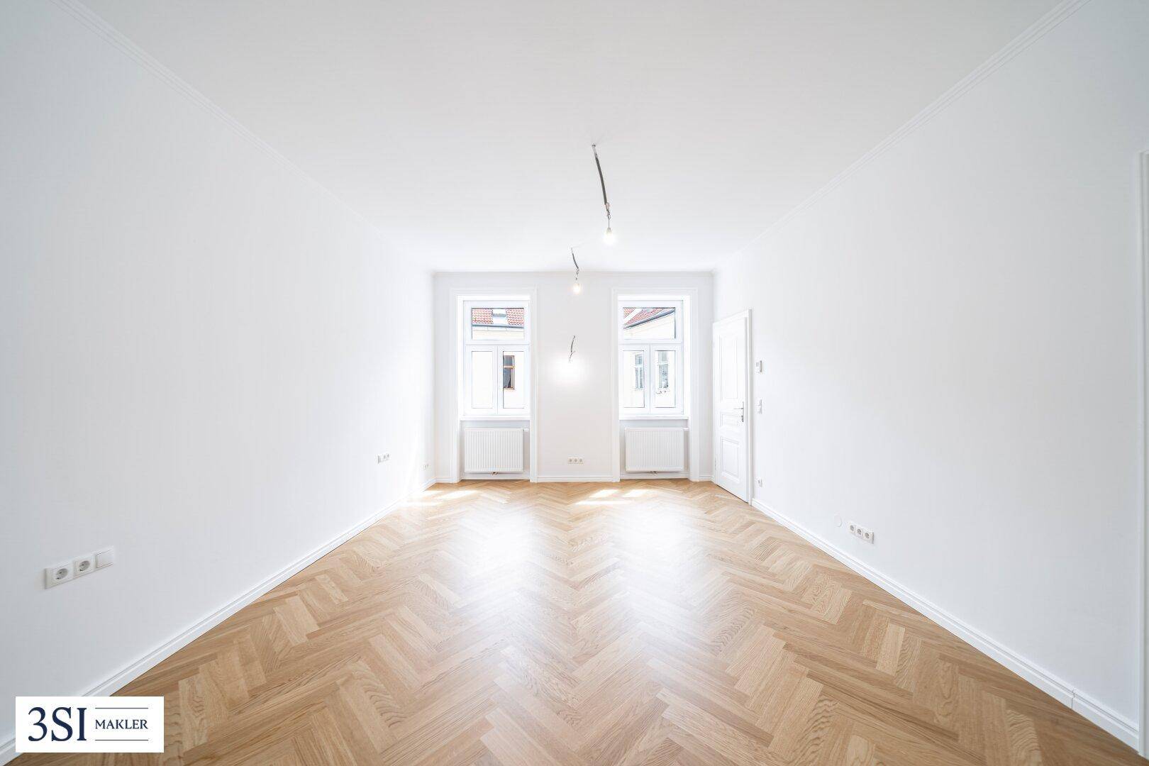 Geräumiges, lichtdurchflutetes Zimmer mit elegantem Fischgrätparkett und weiß gestrichenen Wänden.