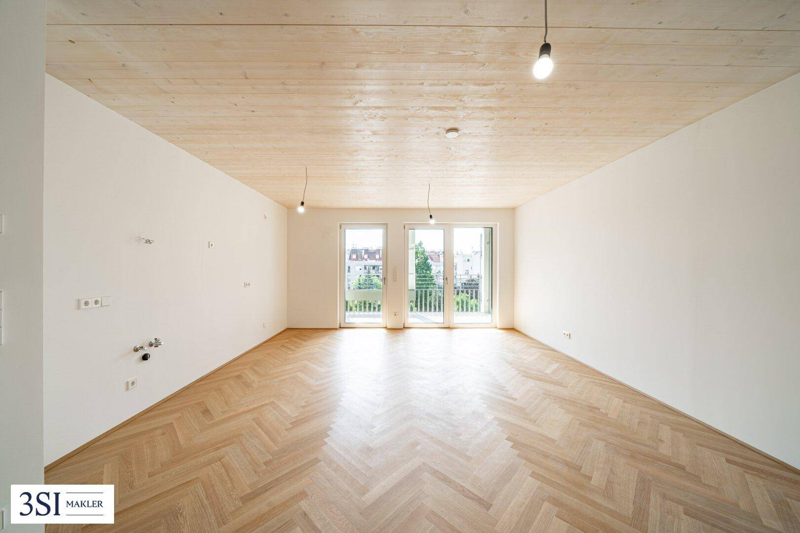 Geräumiges, unmöbliertes Wohnzimmer mit hellem Fischgrätparkett und großen Fenstern zum Außenbereich.