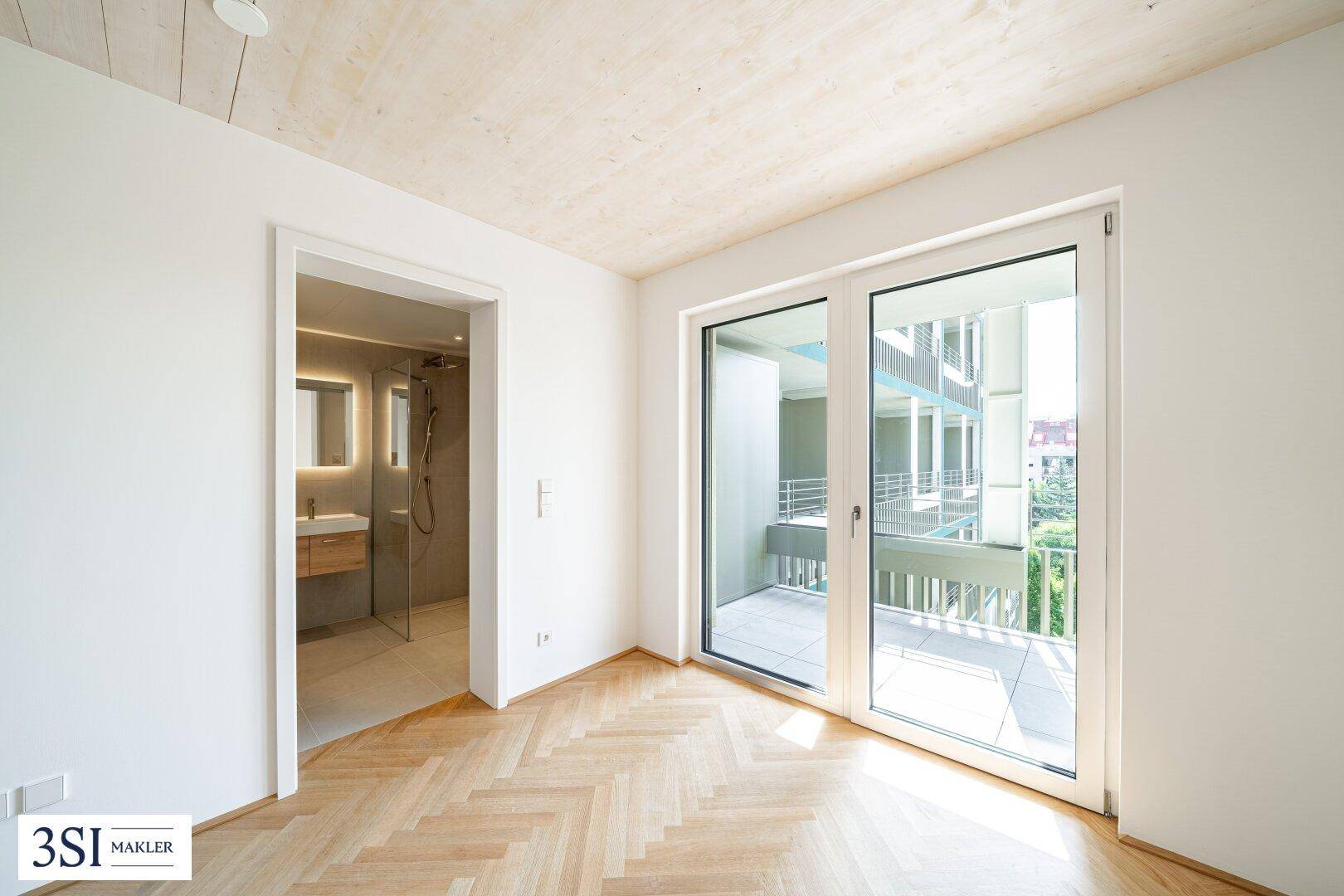 Helles Zimmer mit Fischgrätparkett und Zugang zu einem Balkon sowie einem angrenzenden Badezimmer.