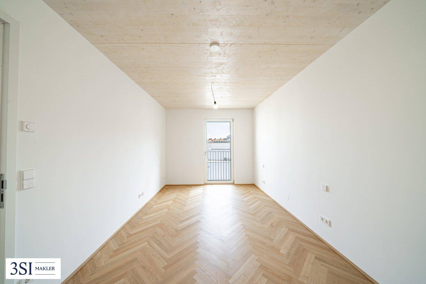 Heller, leerer Raum mit Fischgrätparkettboden, Holzbalkendecke und Zugang zu einem französischen Balkon.