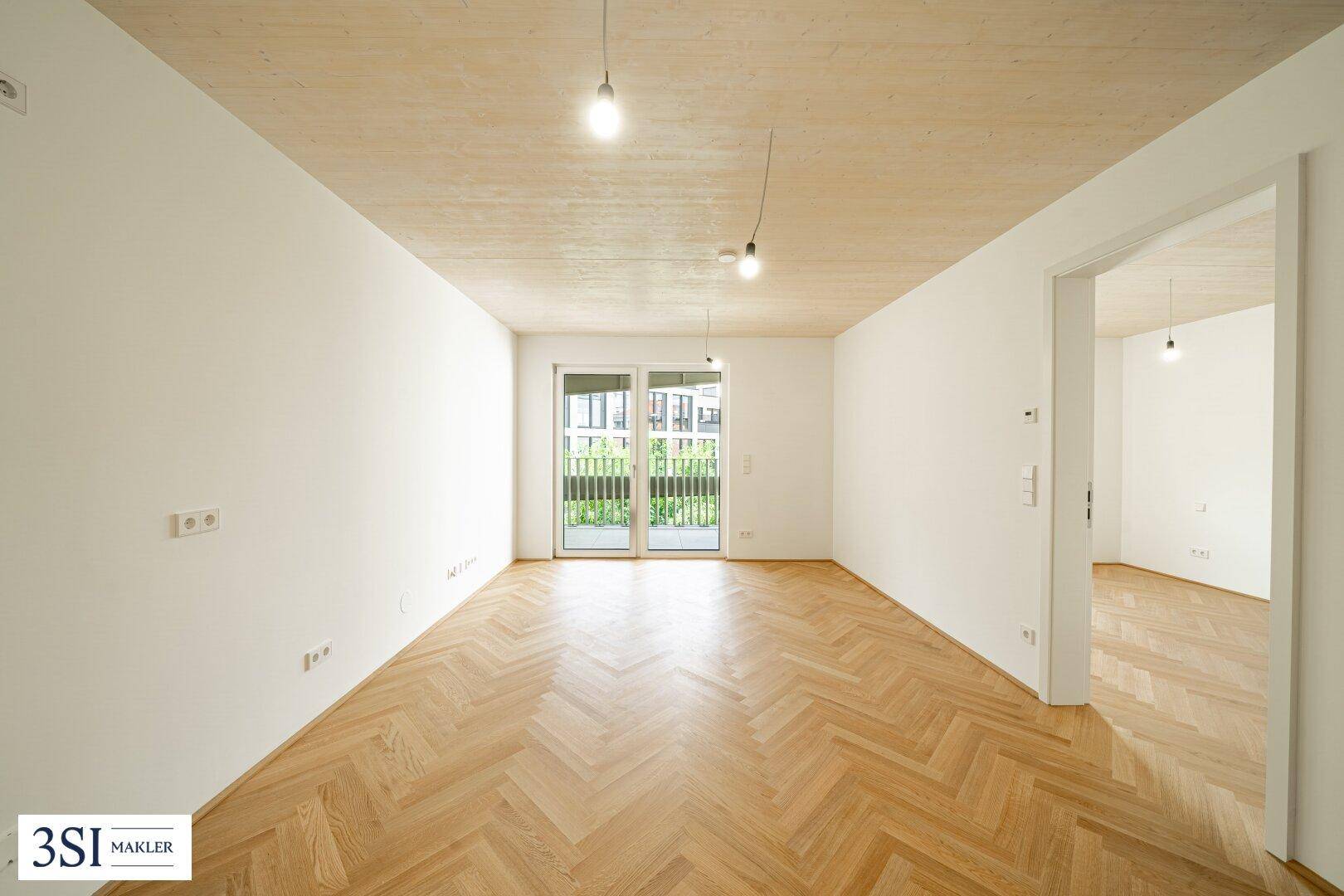 Geräumiger, heller Raum mit Fischgrätparkett, Holzbalkendecke und Zugang zu einem Balkon.