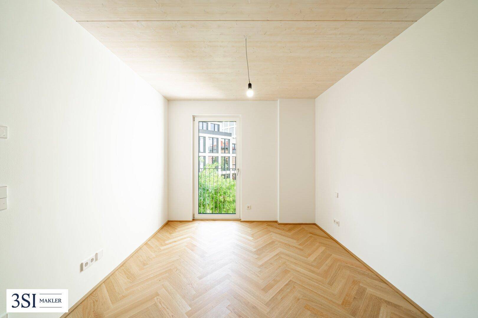 Lichtdurchflutetes Zimmer mit Fischgrätparkett, Holzbalkendecke und einem französischen Balkon mit Grünblick.