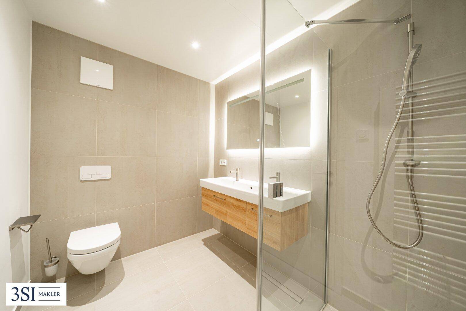 Modernes Badezimmer mit hellgrauen Fliesen, Holzwaschtisch, beleuchtetem Spiegel und einer geräumigen Walk-in-Dusche.