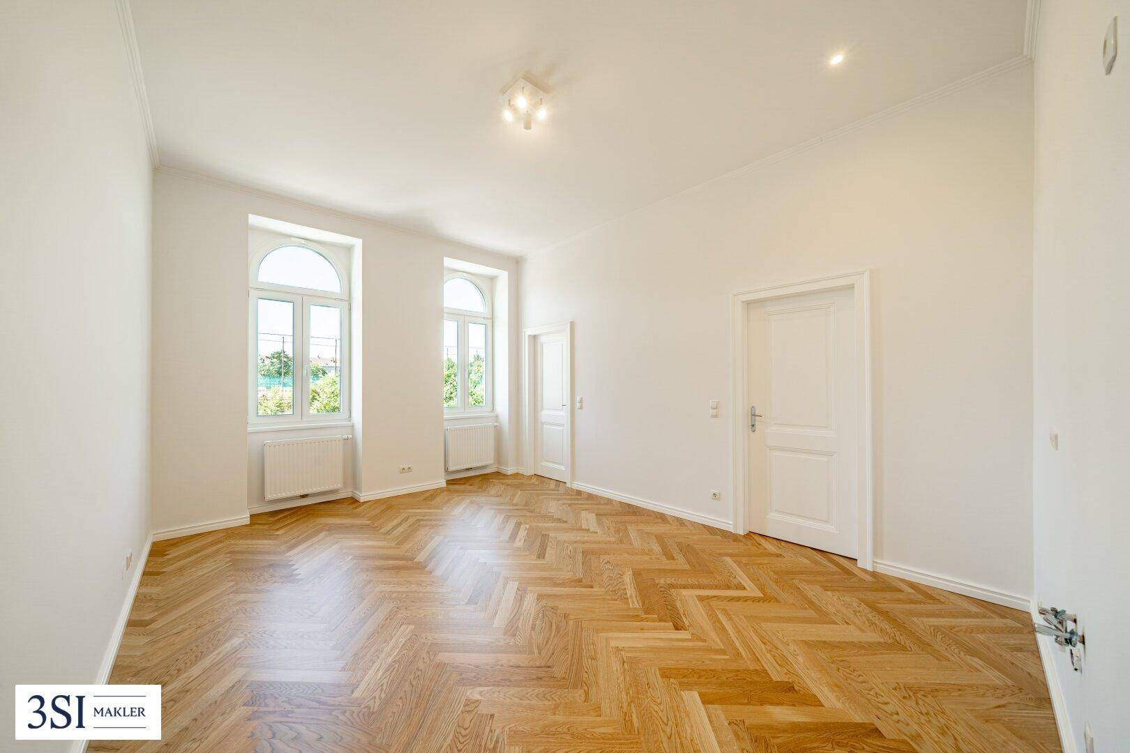 Geräumiges, helles Zimmer mit Fischgrätparkett und hohen Fenstern, ideal als Wohnbereich.