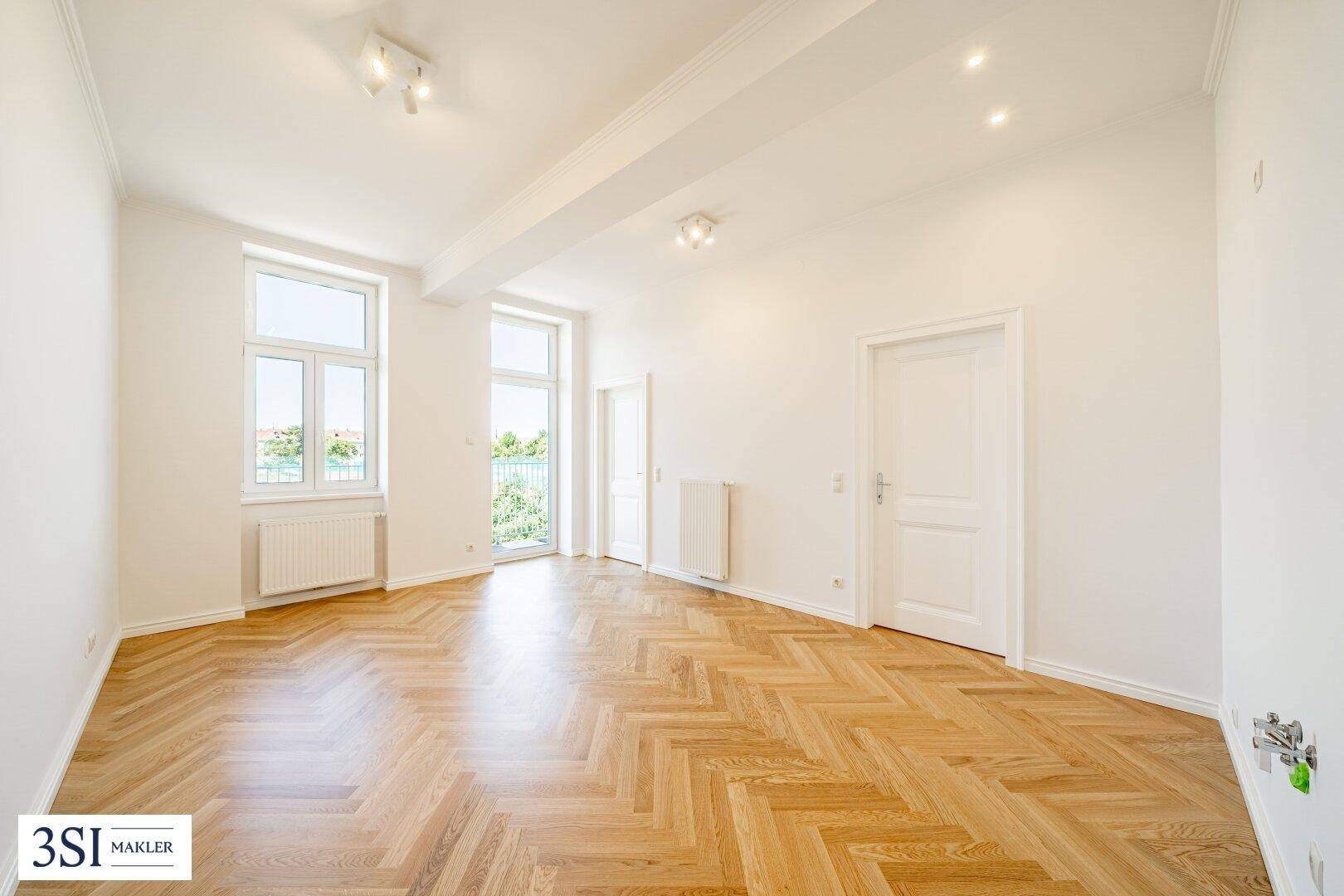 Geräumiger, heller Raum mit Fischgrätparkett und Zugang zu einem Balkon.