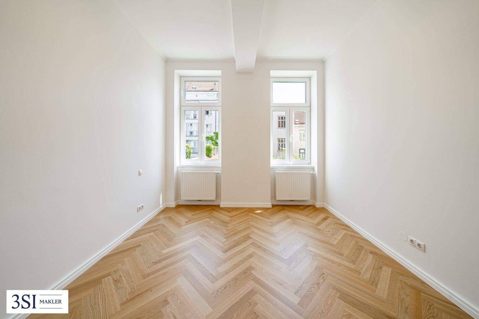 Heller, leerer Raum mit elegantem Fischgrätparkett und zwei großen Fenstern.