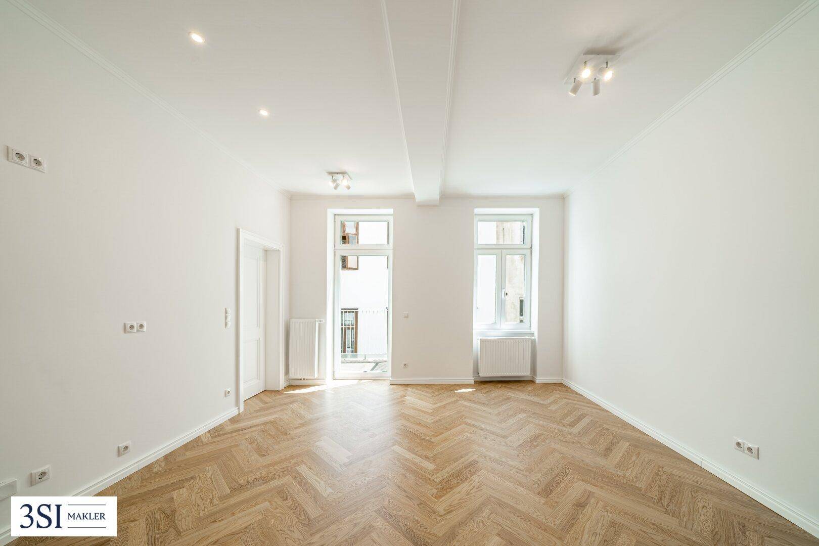Leerer Raum mit Fischgrätparkett und Zugang zu einem französischen Balkon.