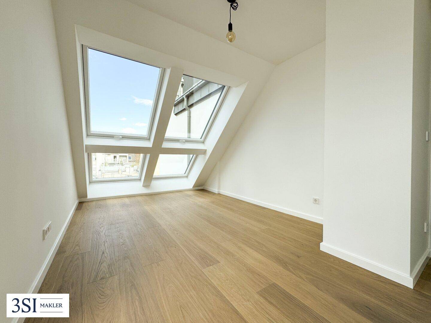Heller Raum mit Holzboden, Dachschrägen und Dachfenstern mit Blick auf die umliegenden Gebäude.