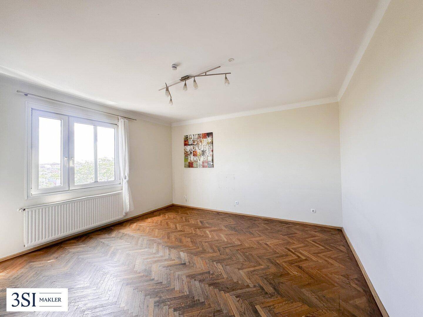 Geräumiges, leeres Zimmer mit Fischgrätparkett und großem Fenster für viel Tageslicht.