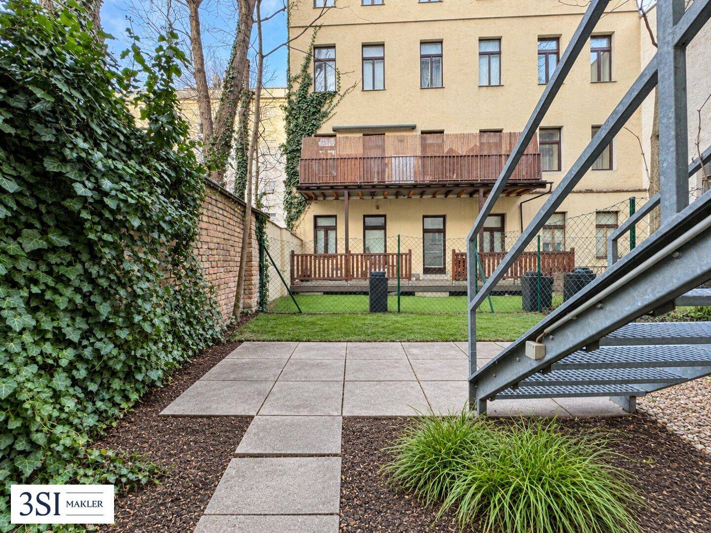 Gepflasterte Terrasse mit Rasenfläche und Efeuwand, angrenzend an ein Gebäude mit Balkonen.