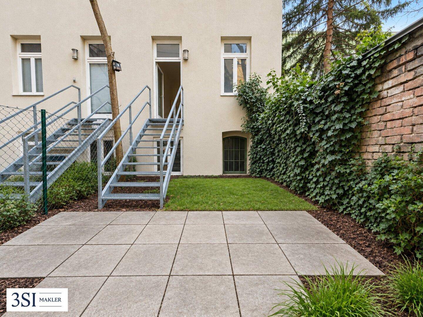 Sonniger Gartenbereich mit gepflasterter Terrasse, Rasen und Zugang zum Gebäude über eine Metalltreppe.