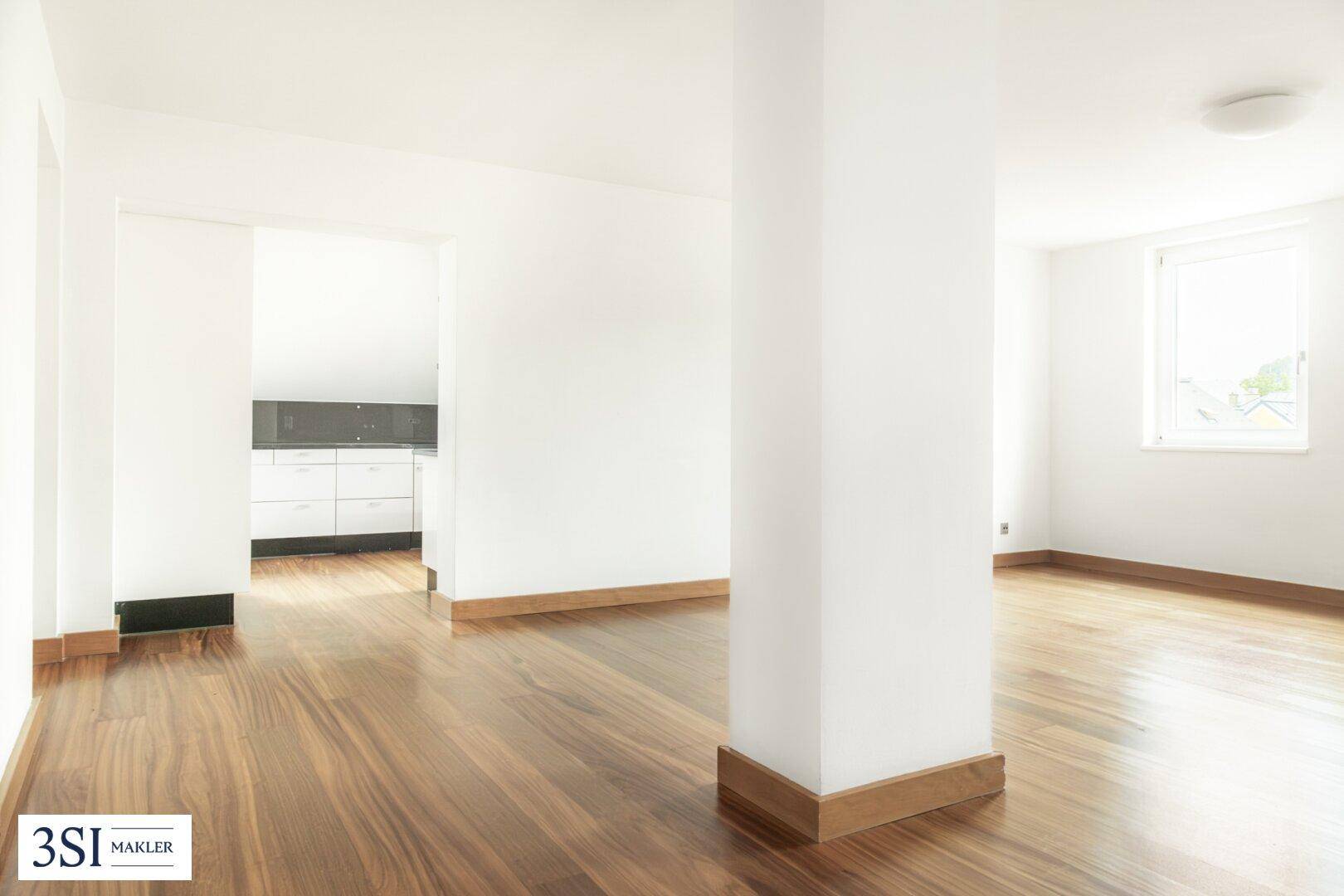 Geräumiges, helles Wohnzimmer mit Holzboden und Blick auf den angrenzenden Küchenbereich.