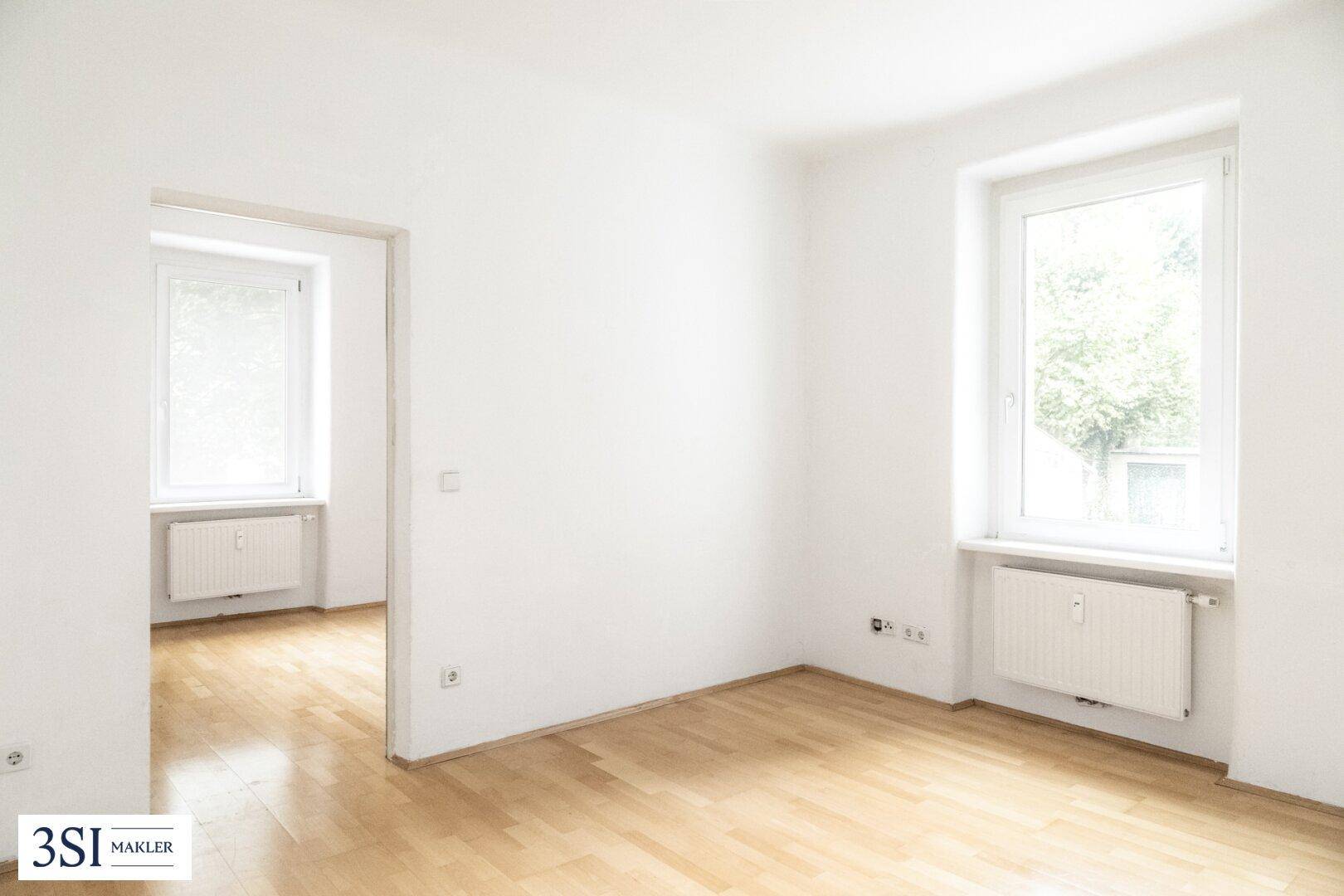 Leerer Wohnraum mit hellem Parkettboden und großen Fenstern.