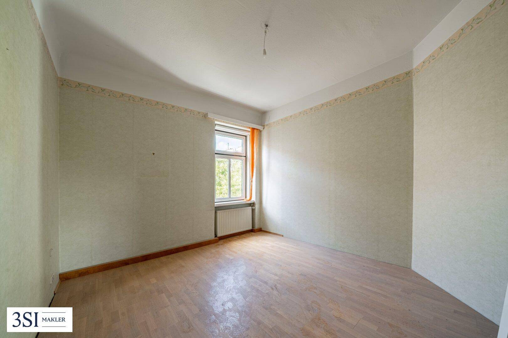 Schlichter Raum mit hellem Parkettboden und einem Fenster, das viel Tageslicht hereinlässt.
