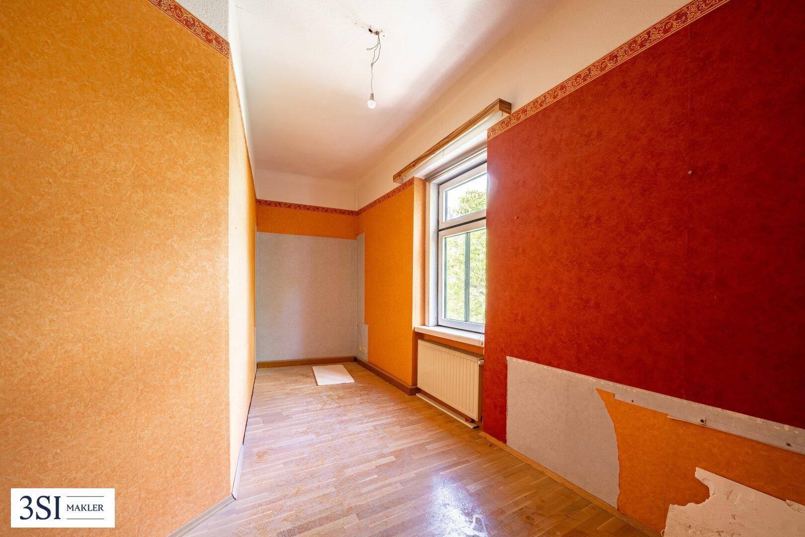 Zimmer mit auffälliger orange-roter Tapete, Parkettboden und einem Fenster, das Licht spendet.