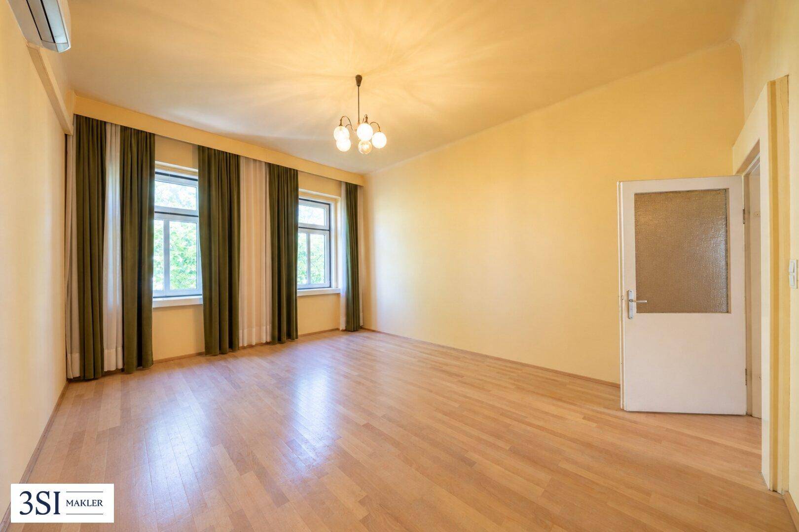 Geräumiges Zimmer mit Holzparkett, gelben Wänden und zwei Fenstern mit Vorhängen.