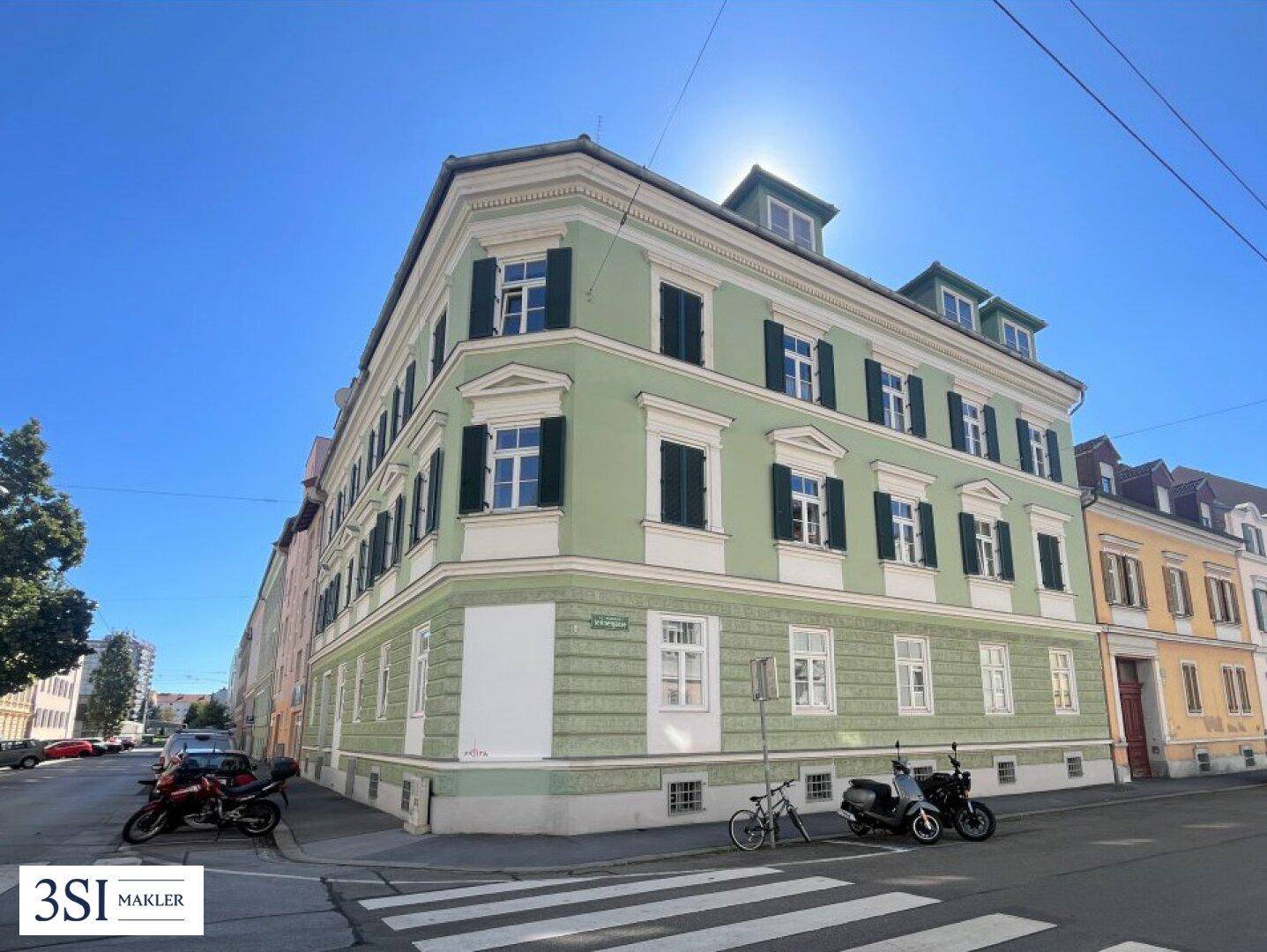 Historisches Eckgebäude mit grüner Fassade, weißen Fensterrahmen und traditionellen Fensterläden unter blauem Himmel.