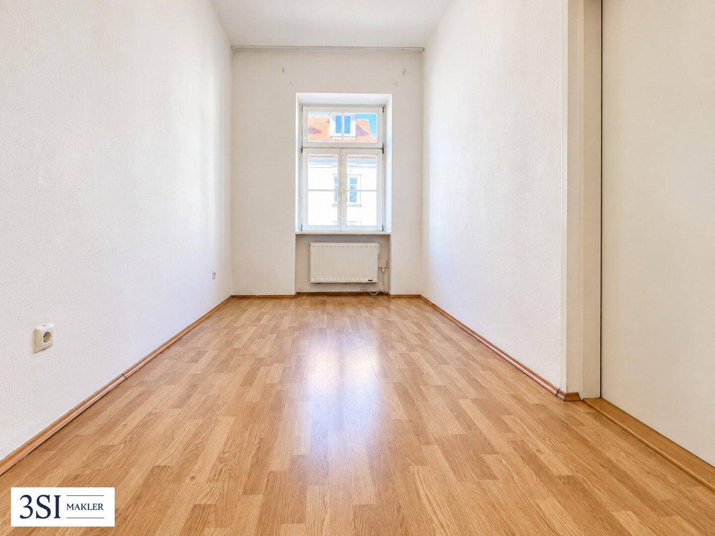 Leerer Raum mit Laminatboden, einem Fenster und Heizkörper, ideal zur individuellen Gestaltung.