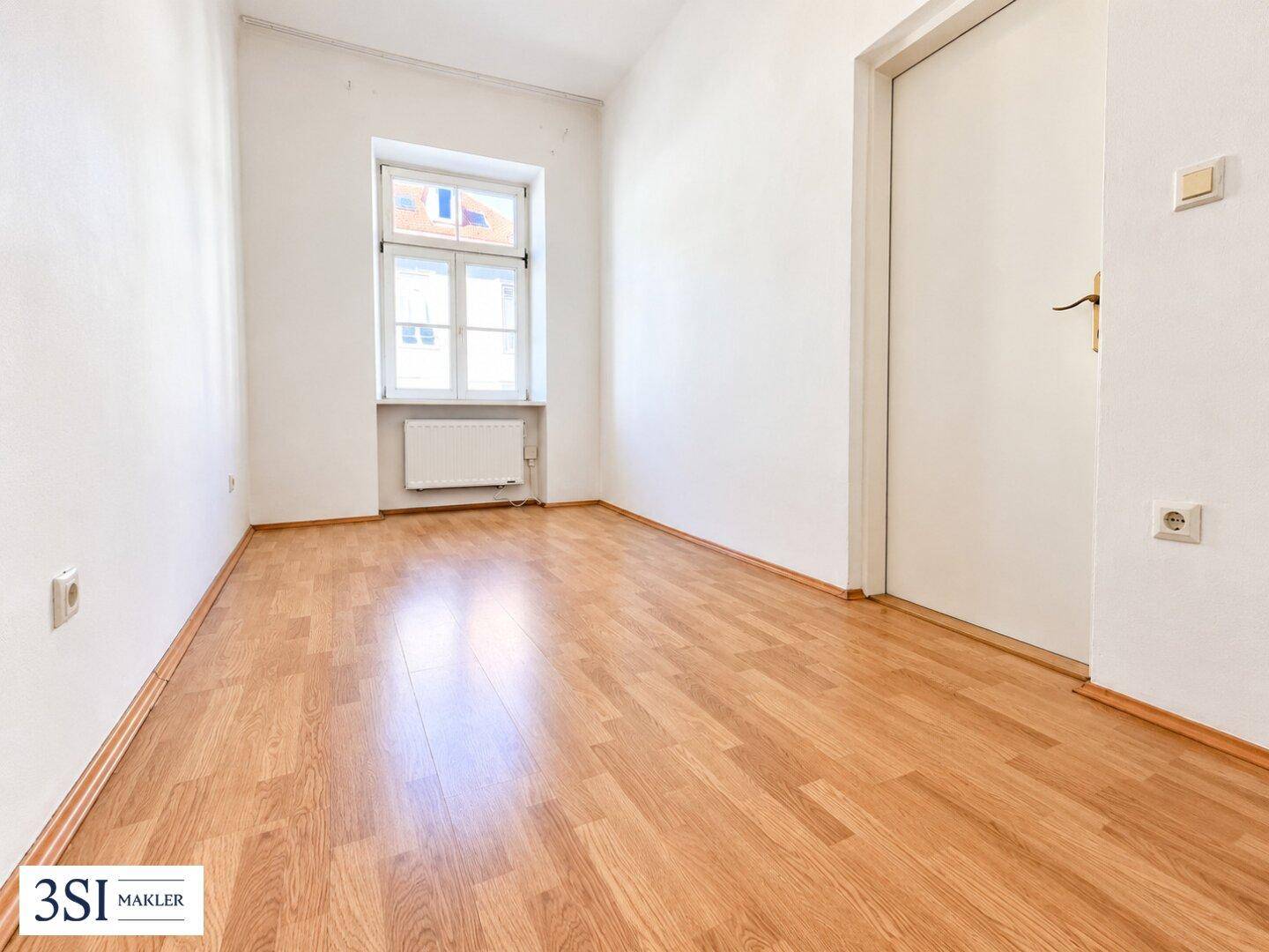 Heller, leerer Raum mit Laminatboden, Fenster und weißer Tür, bereit zum Einzug.