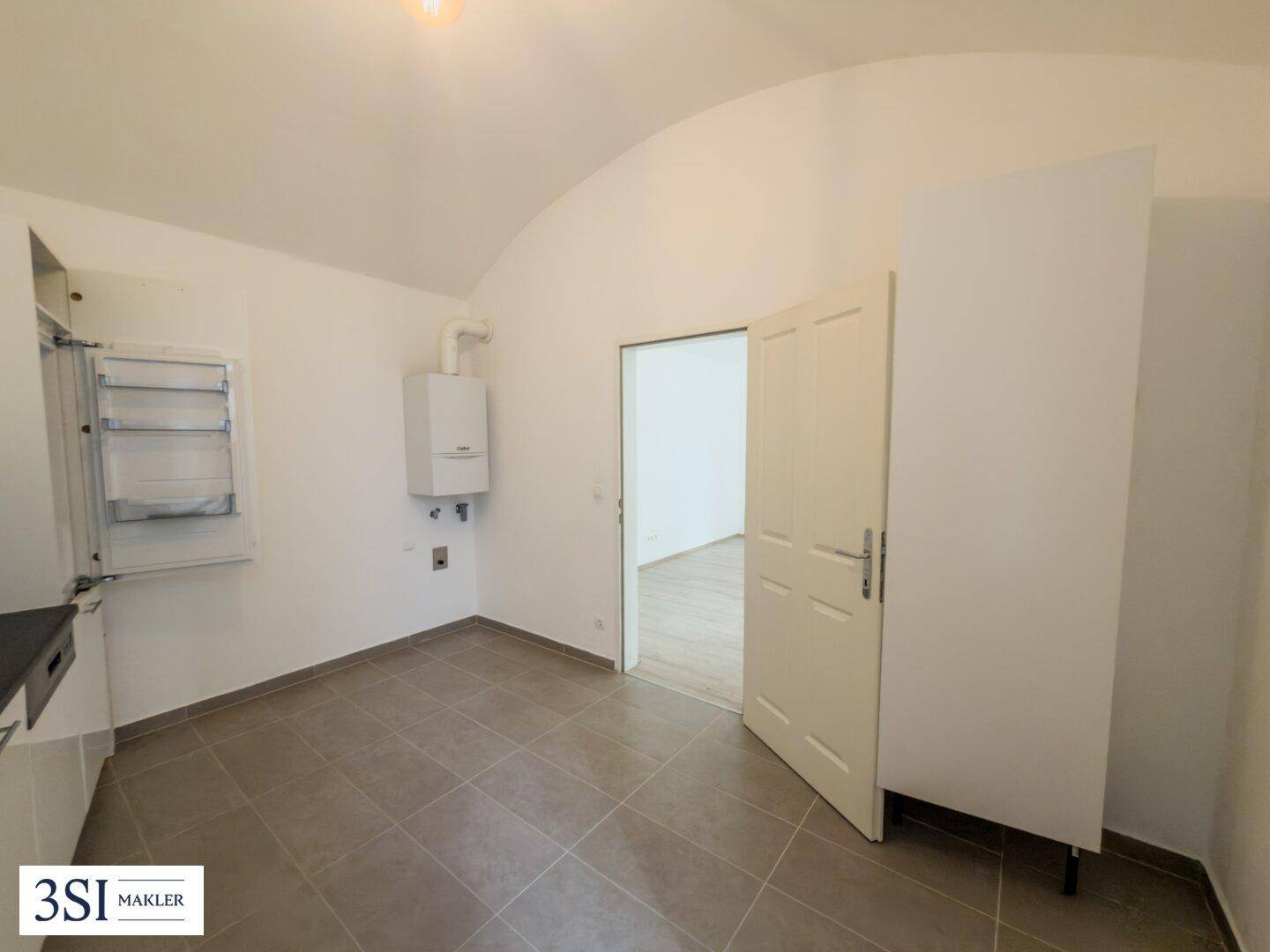 Küchenbereich mit Fliesenboden, einem Warmwasserbereiter an der Wand und Zugang zu einem weiteren Raum.