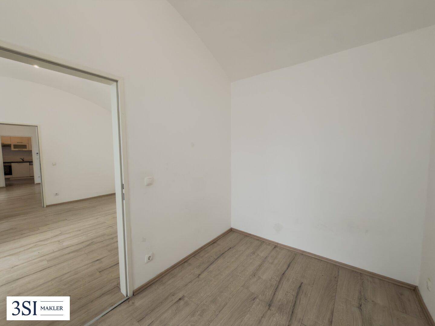 Leeres Zimmer mit Laminatboden und weiß gestrichenen Wänden, bietet Zugang zu einem angrenzenden Küchenbereich.