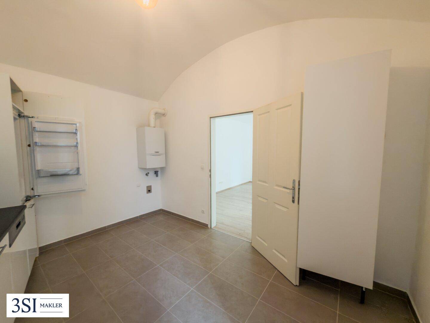 Küchenbereich mit Fliesenboden, einem Warmwasserbereiter an der Wand und Zugang zu einem weiteren Raum.