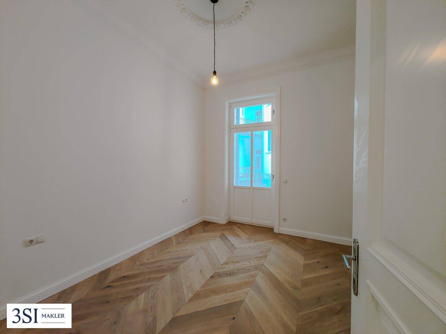 Helles Zimmer mit Fischgrätparkett, Stuckdecke und Zugang zu einem möglichen Balkon oder Austritt.