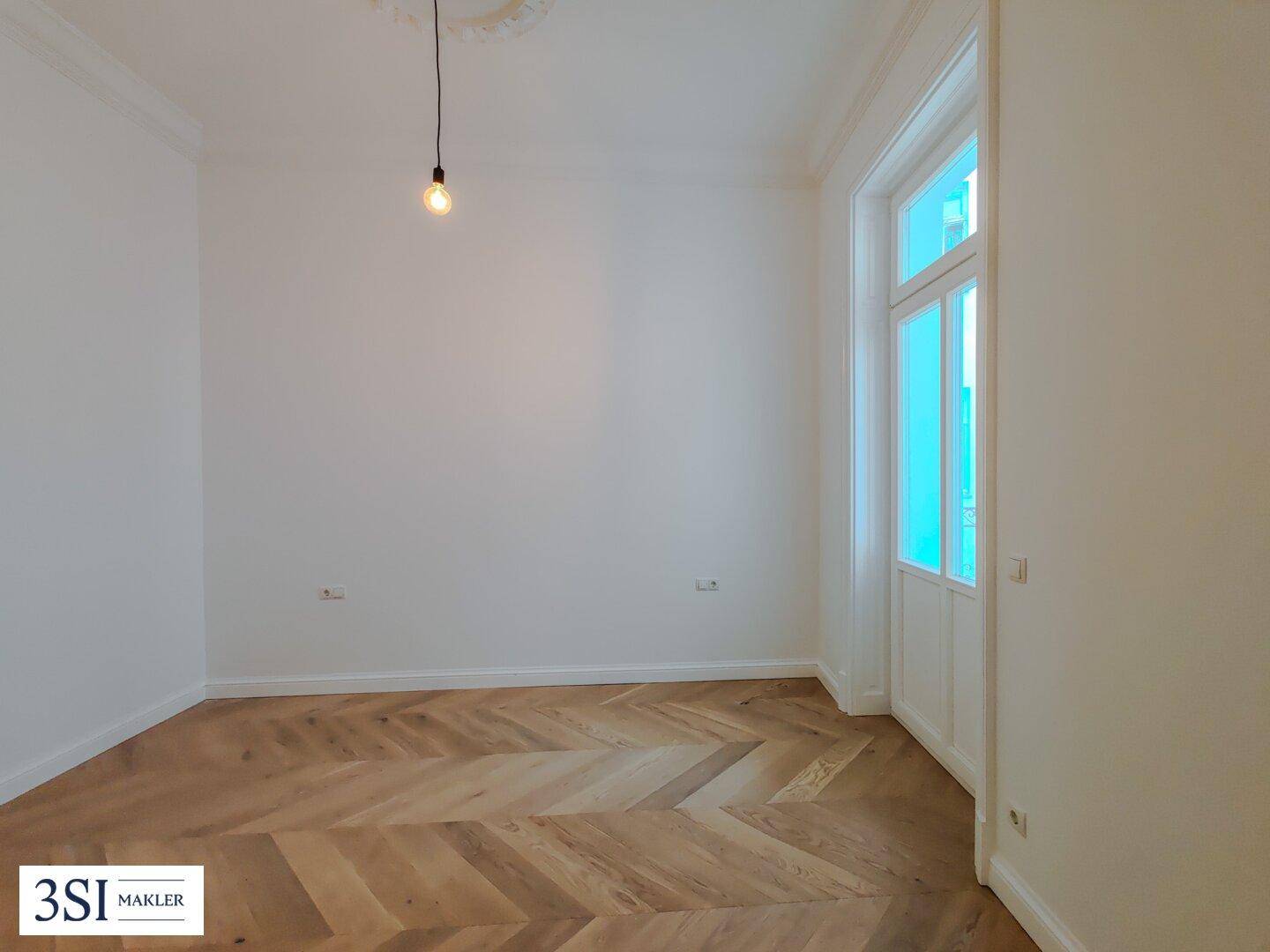 Geräumiges Zimmer mit Fischgrätparkett und großen Fenstern, ideal für viel Tageslicht und frische Luft.