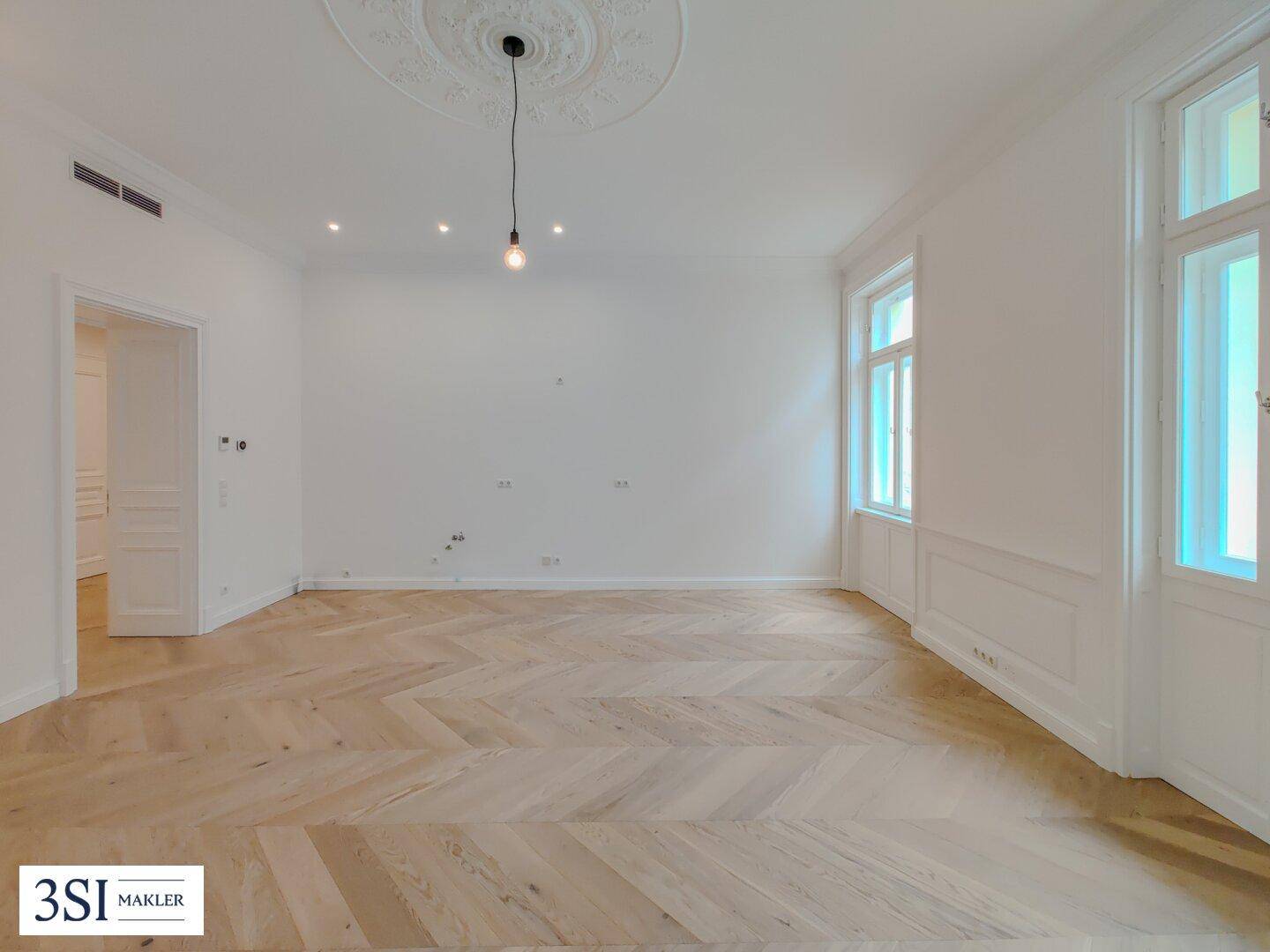 Geräumiges Zimmer mit elegantem Fischgrätparkett, Stuckdecke und viel Tageslicht durch große Fenster.