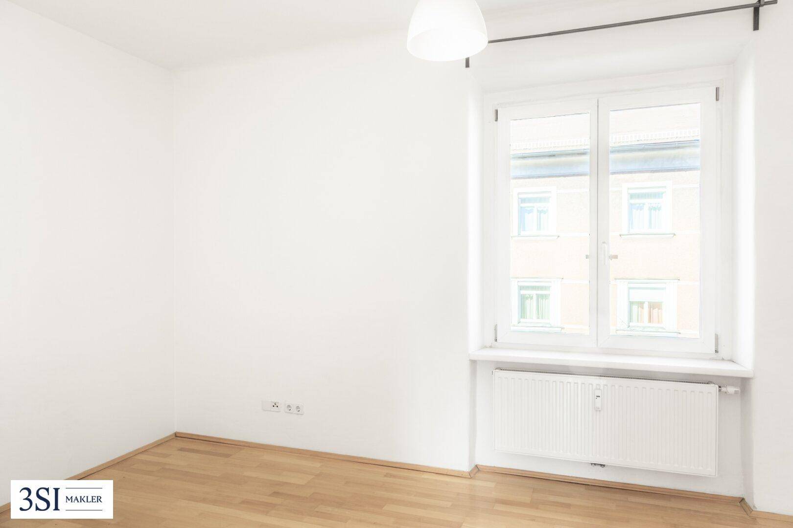 Leeres Zimmer mit hellem Holzfußboden, weißen Wänden und einem großen Fenster für viel Tageslicht.
