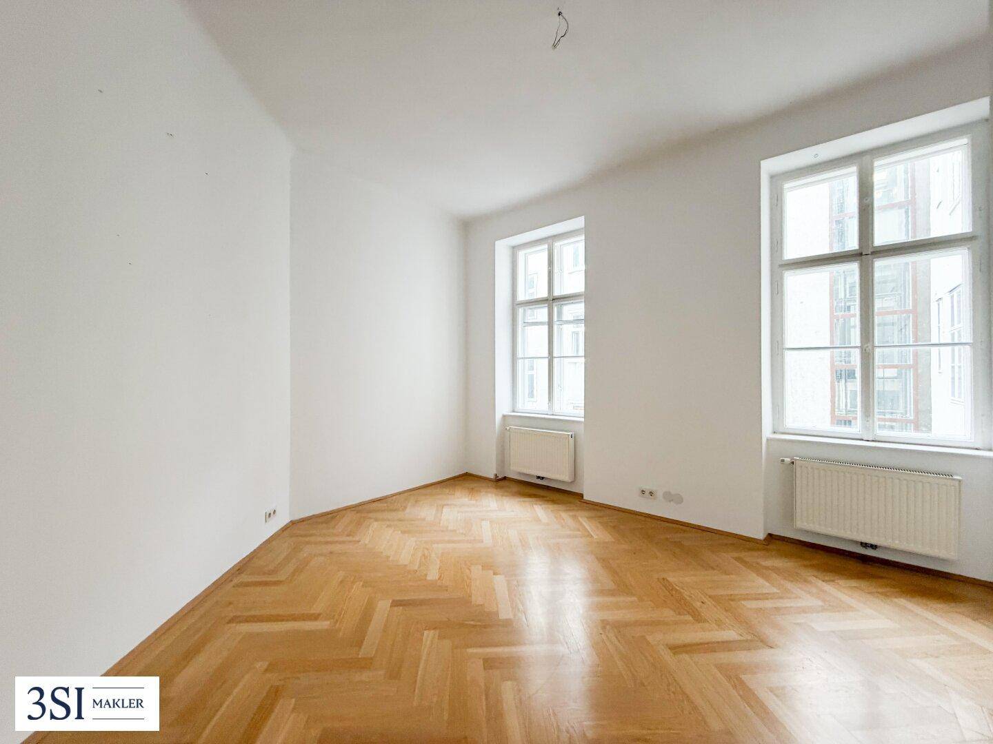 Heller, leerer Raum mit Fischgrätparkett und hohen Fenstern, ideal als Schlaf- oder Arbeitszimmer.