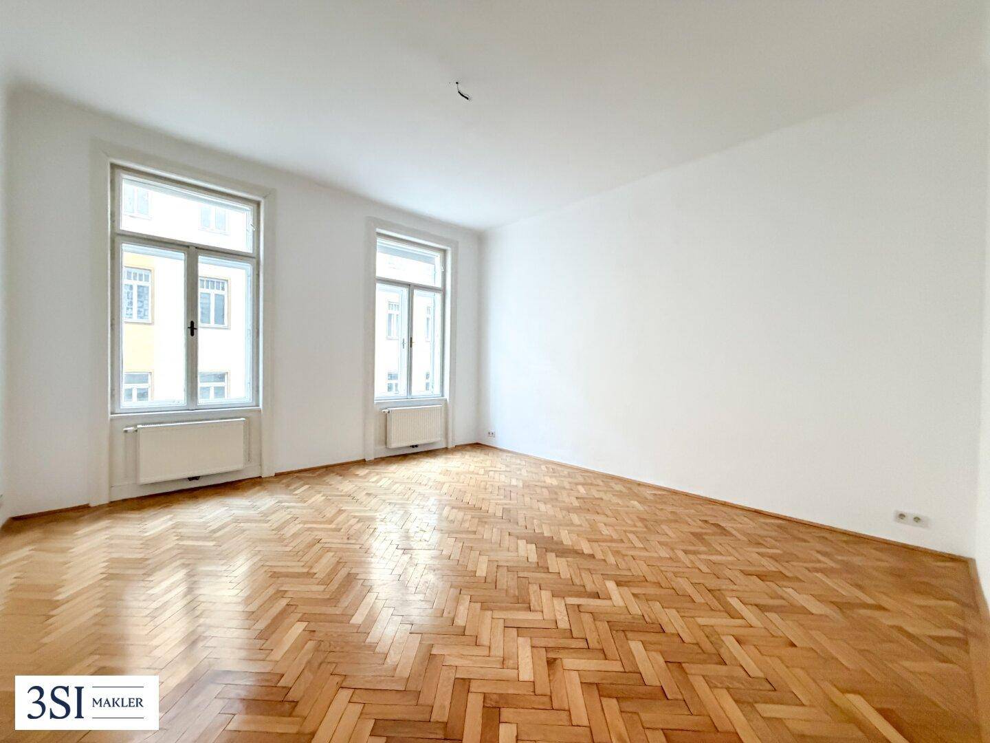 Geräumiger, unmöblierter Raum mit Fischgrätparkett und großen Fenstern, bereit zur individuellen Gestaltung.