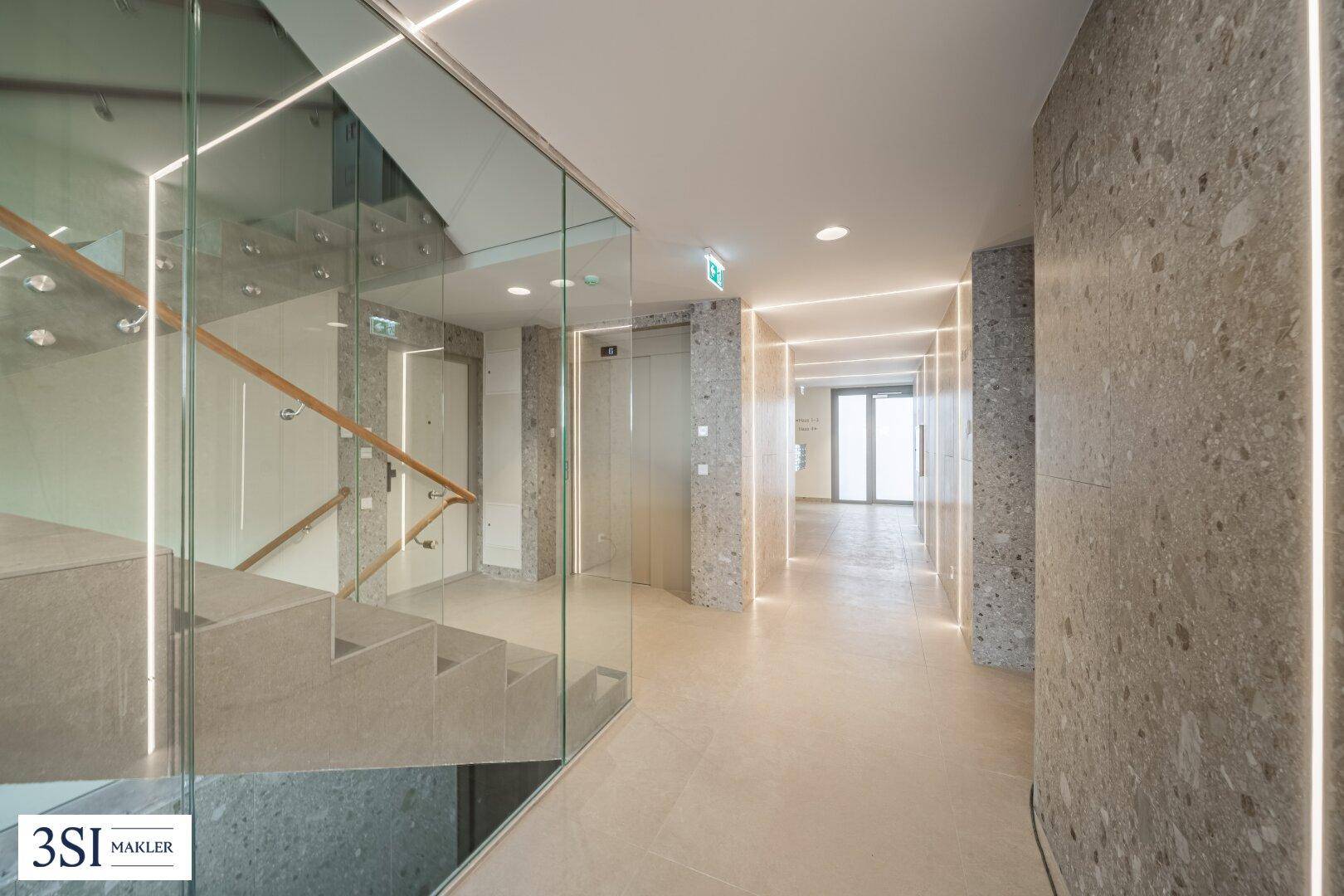 Heller Flur mit Treppenhaus, Glasgeländer und Aufzug in modernem Design und Steinwänden.