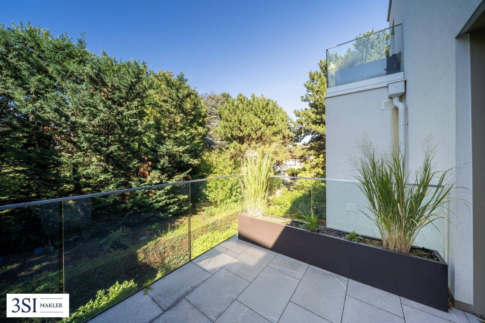 Moderner Balkon mit Glasgeländer, dekorativen Pflanzen und Blick ins Grüne.