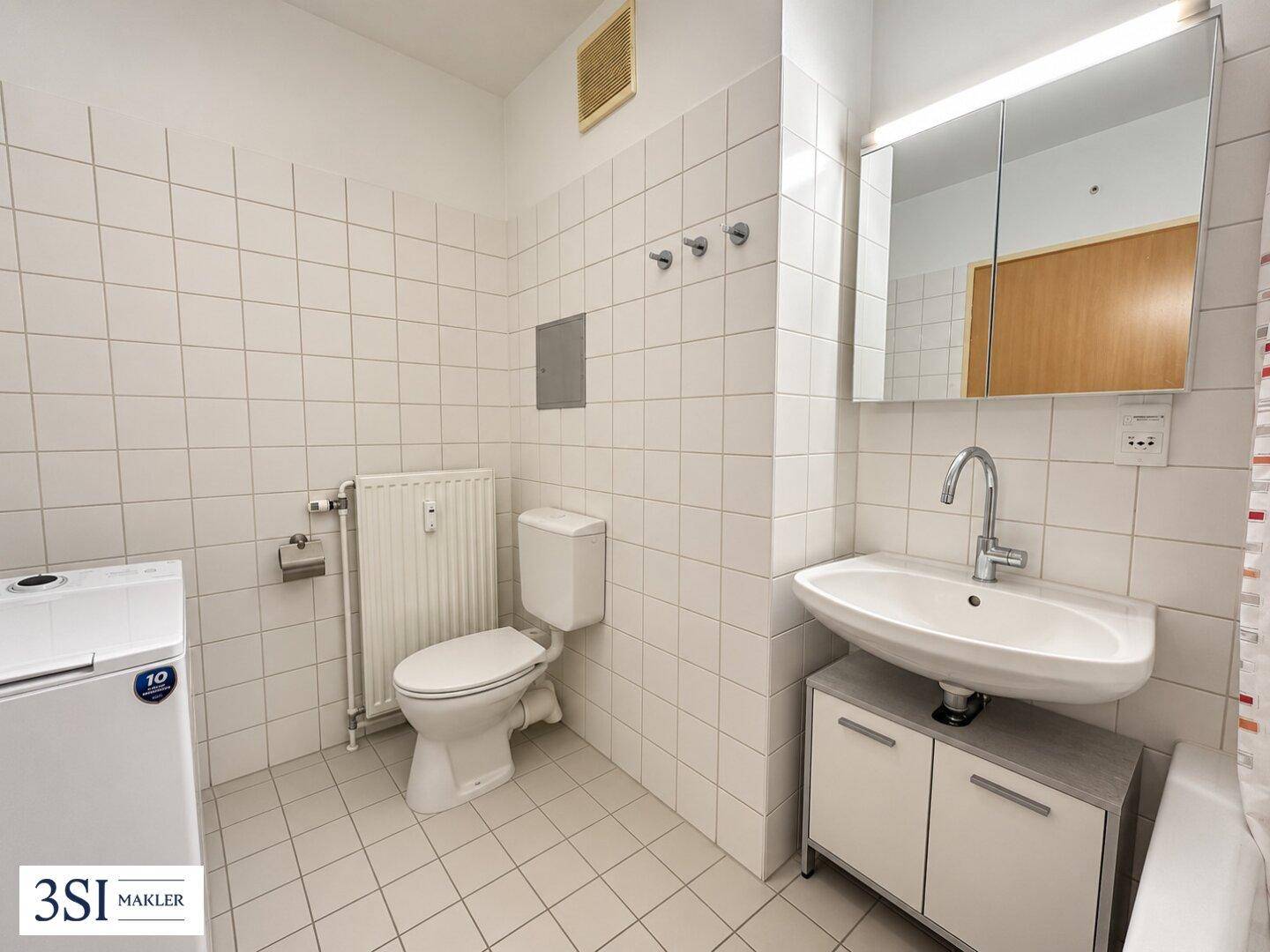 Helles Badezimmer mit weißen Fliesen, WC, Waschbecken, Spiegelschrank und einer Waschmaschine.
