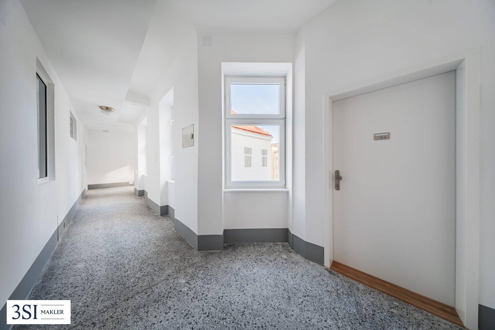 Heller, langer Flur mit Fenstern und einem robusten Terrazzoboden in einem Mehrfamilienhaus.