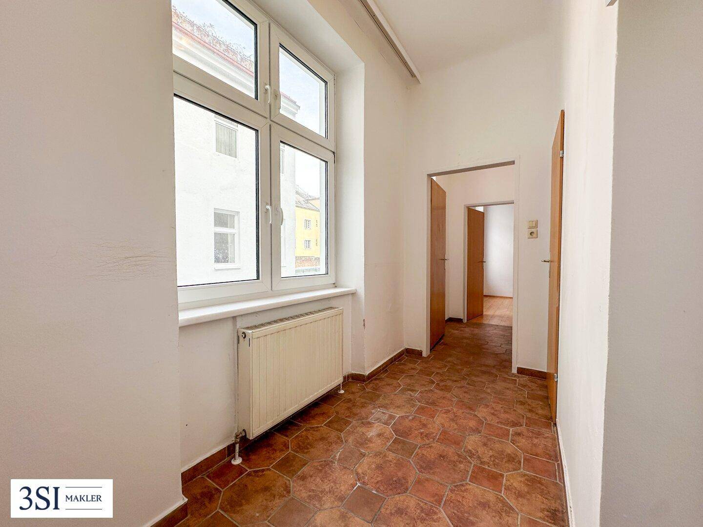 Heller Flur mit Terrakotta-Fliesenboden und einem großen Fenster, das Tageslicht hereinlässt.