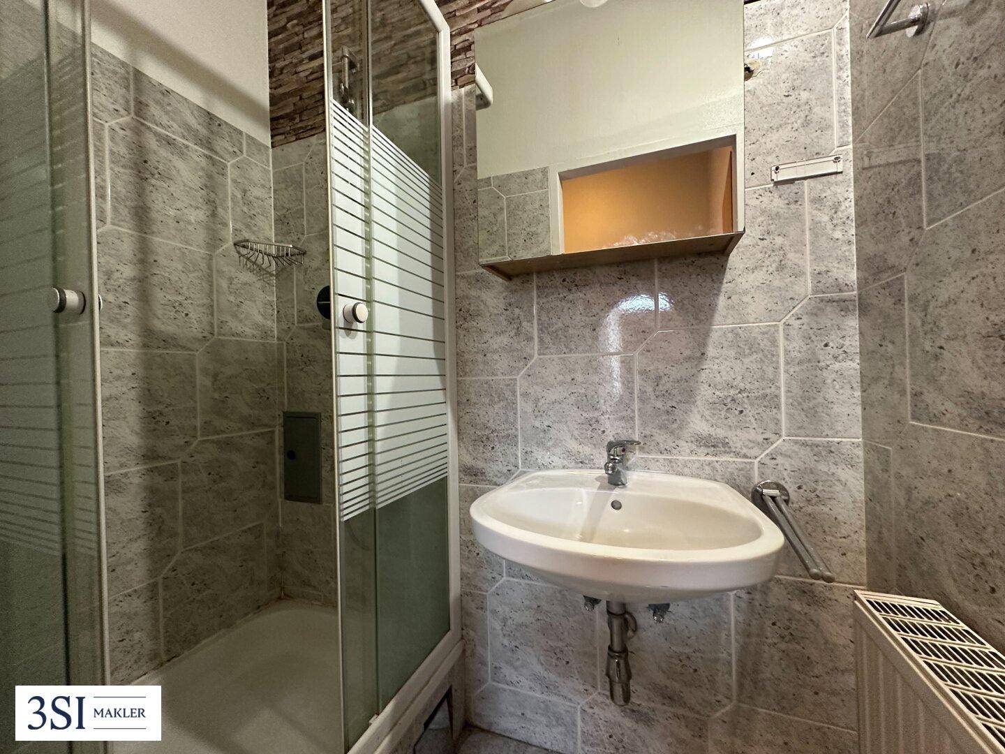 Kleines Badezimmer mit Duschkabine, Waschbecken und grauen Fliesen an Wänden und Boden.
