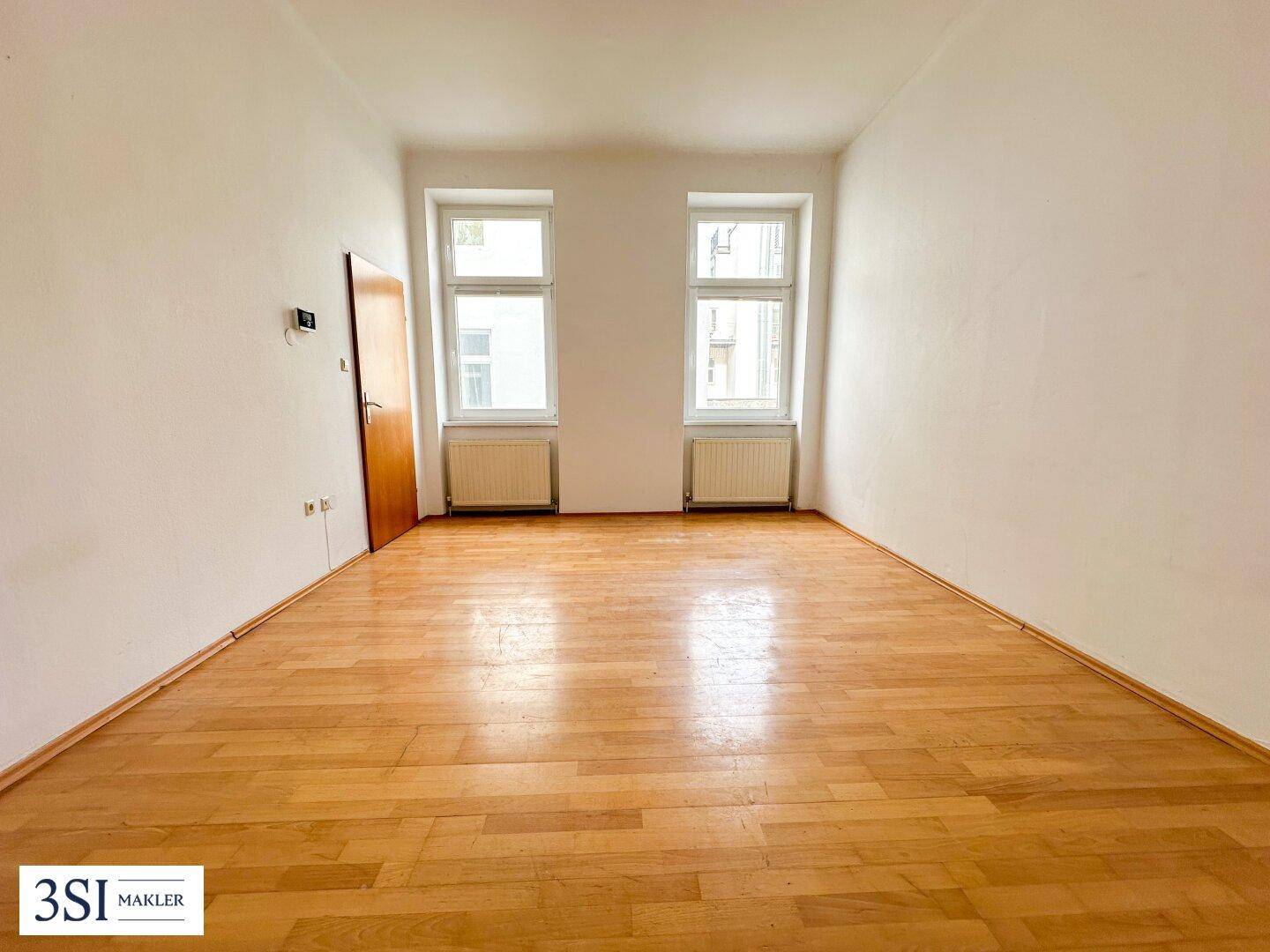 Leeres Zimmer mit hellem Holzboden und zwei Fenstern, ideal für Wohn- oder Schlafbereich.